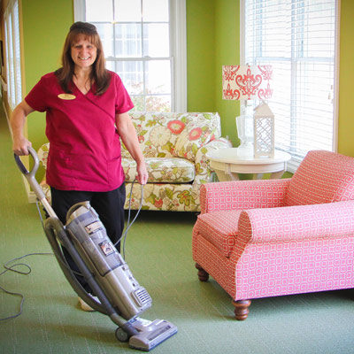 A woman that is working as a house keeper is holding a vacume machine wearing pink.