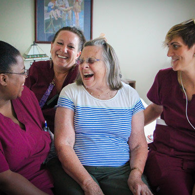 A Woman wearing a white and blue shirt laughing with three other women wearing the same red uniform working in the same senior care career